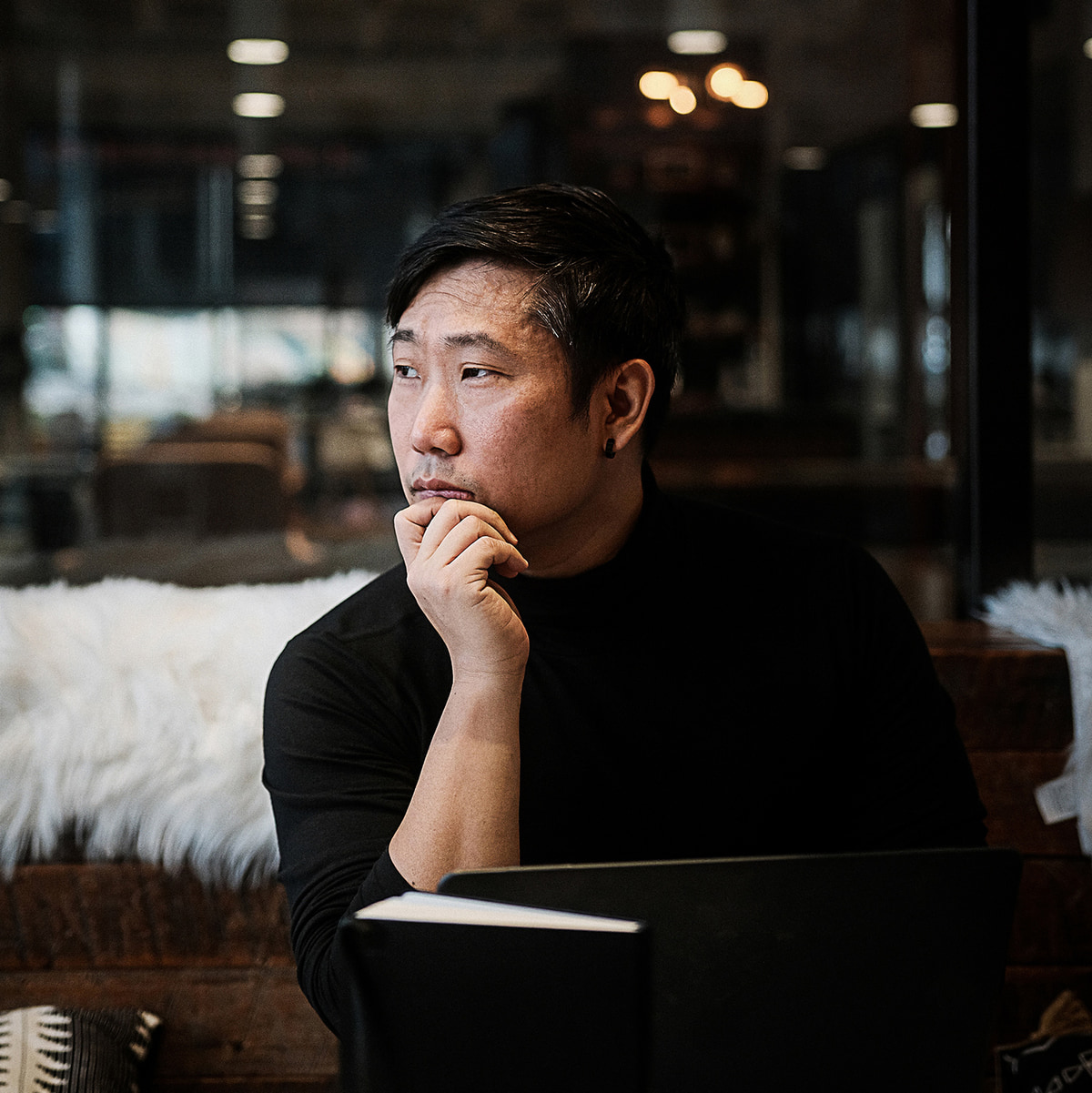 Won J. You in a black turtleneck, seated at a laptop with his chin resting on his hand, looking away in thought in a dimly lit studio